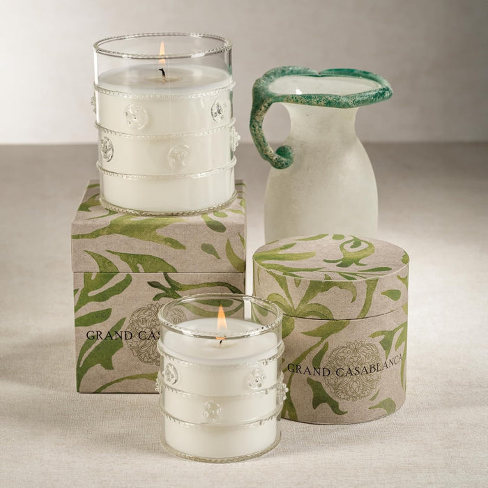 The ZODAX GRAND CASABLANCA CANDLE - WILD TUBEROSE, 6.5OZ features white wax in a jar beside a round box with green leafy patterns, filling the air with wild tuberose—a beautiful botanical gift candle for any occasion.