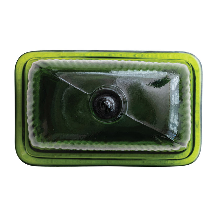 The CREATIVE COOP GREEN PRESSED GLASS BUTTER DISH is displayed with green glass containers and bottles on a textured white surface, beside a folded towel and a partially visible book.