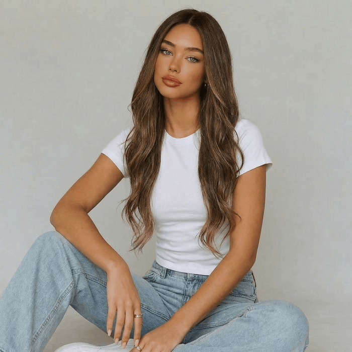 Model wearing a long highlighted brown body wave wig with soft waves, dressed in a white t-shirt and blue jeans.