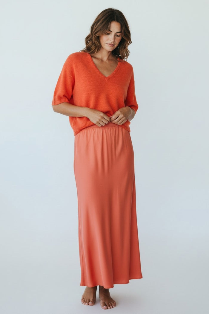 A woman stands barefoot against a plain background, wearing an orange V-neck short-sleeve knit top and the ROSE & CROWN SATIN MIDI SKIRT by ROSE & CROWN, featuring a coral hue and elastic waistband. She looks down, gently holding her skirt's waistband.