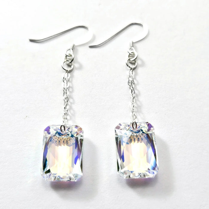 The VERDIER - SWAROVSKI EMERALD CUT CRYSTAL EARRINGS by VERDIER feature sterling silver hooks and rectangular, emerald cut crystals inspired by the Swarovski Aurora design, delicately suspended from fine chains and elegantly presented against a white background.