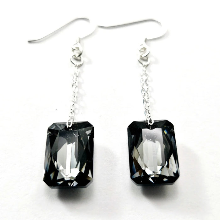 Introducing the VERDIER - SWAROVSKI NIGHT CRYSTAL EARRINGS by VERDIER: a pair of sterling silver drop earrings featuring rectangular, emerald-cut Swarovski crystals suspended from delicate chains, beautifully showcased against a white background.