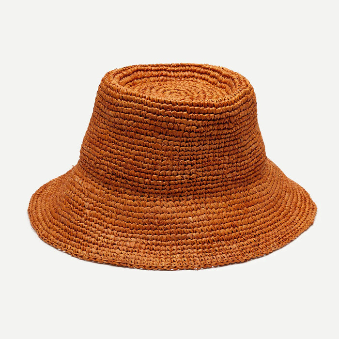 A woman with long, wavy blonde hair wears the WYETH TALI Raffia Bucket Hat in Rust and a white sleeveless top, standing in profile against a plain light background.