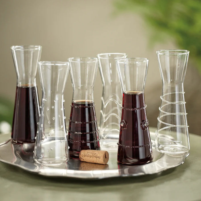 Six ZALLI SINGLE SERVE GLASS CARAFES by ZODAX, each featuring a unique etched design, are arranged on a reflective silver tray with a blurred green background.