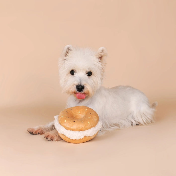 A small, fluffy white dog sniffs the CHEWFFON BAGEL PLUSH SQUEAKY DOG TOY, a large, round, beige plush toy resembling a sandwich, set against a plain light beige background.