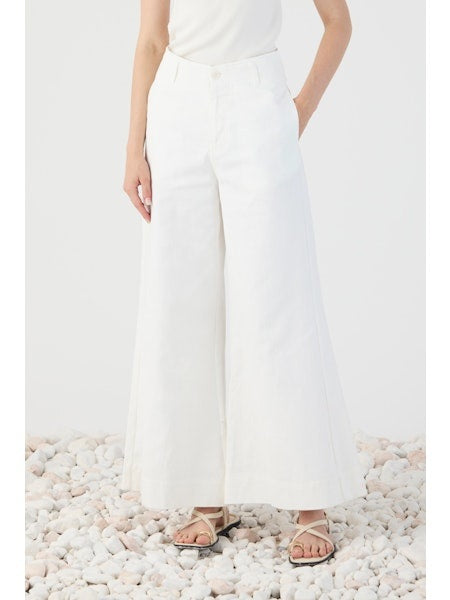 A person stands on white pebbles, shown from the back, wearing ELEH’s WIDE LEG COTTON TROUSER paired with a white sleeveless top and sandals—an effortless take on modern minimalism.