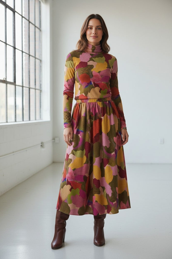 A woman stands indoors wearing the FRANCO FERRARI FRAGOLINA TURTLENECK, a long-sleeve dress featuring a bold abstract floral and leaf pattern in vibrant red, pink, yellow, and green. She has wavy brown hair and smiles softly at the camera.