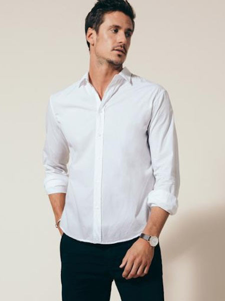 A man in the FRANK & EILEEN Finbar Men's Button Up Shirt in classic white poplin stands with rolled sleeves and hands in pockets, looking to the side. He has short dark hair, wears a watch, and poses against a light background.