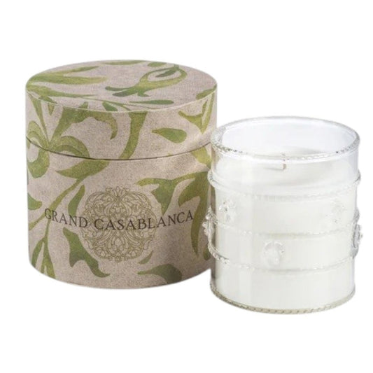 Two lit ZODAX Grand Casablanca Candle – Wild Tuberose, 6.5oz, in decorative glass holders rest on patterned boxes beside a frosted white pitcher with a green rim, all on a neutral surface, evoking the wild tuberose scent.