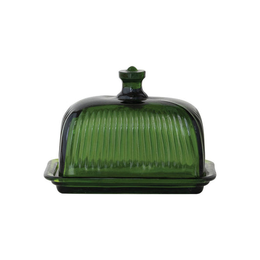 The CREATIVE COOP GREEN PRESSED GLASS BUTTER DISH is displayed with green glass containers and bottles on a textured white surface, beside a folded towel and a partially visible book.