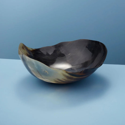 The HORN WAVY ROUND BOWL by BE HOME, INC is an ethically handmade, irregularly shaped bowl crafted from dark marbled horn in Vietnam. It features smooth edges and a natural glossy finish, shown resting on a light blue surface against a blue background.