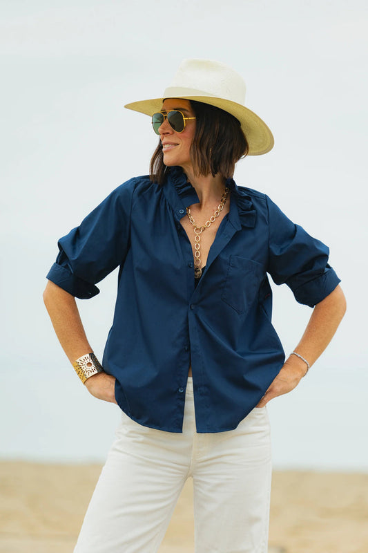 A person stands outdoors with hands on hips, smiling, wearing sunglasses, a wide-brimmed hat, gold jewelry, white pants, and the KMJ SOUTHERN LOVER SHIRT IN NAVY by KMJ. The background is light and neutral.