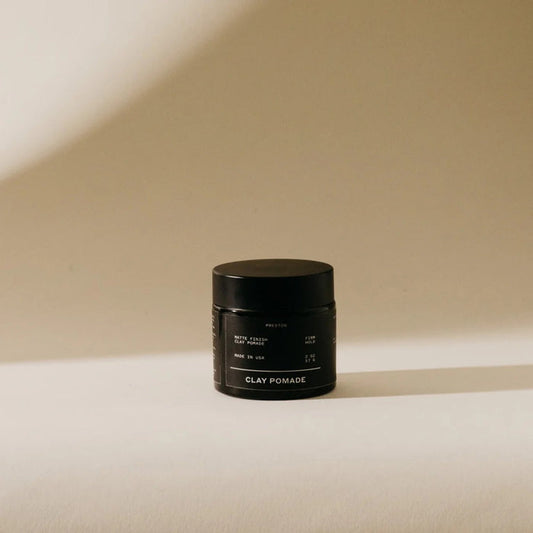A black jar labeled "PRESTON - CLAY POMADE UNSCENTED" from PRESTON, a matte hair clay, sits on a beige surface with a soft shadow on the wall behind it under warm lighting.