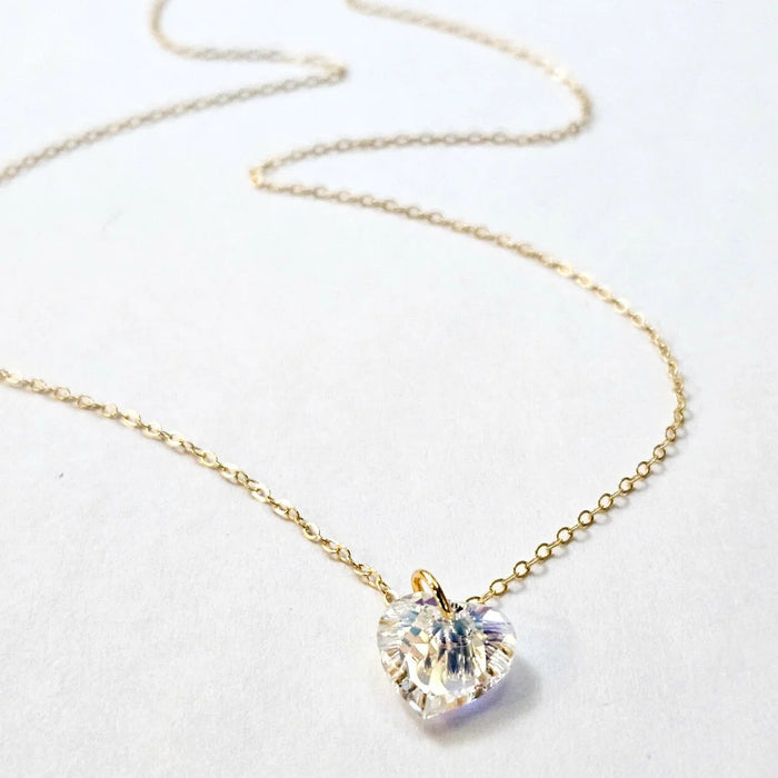 The VERDIER - AURORA CRYSTAL HEART NECKLACE from VERDIER features a sparkling crystal AB heart-shaped pendant suspended on a delicate 14k gold filled chain, shimmering against a white background.