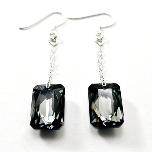 Introducing the VERDIER - SWAROVSKI NIGHT CRYSTAL EARRINGS by VERDIER: a pair of sterling silver drop earrings featuring rectangular, emerald-cut Swarovski crystals suspended from delicate chains, beautifully showcased against a white background.