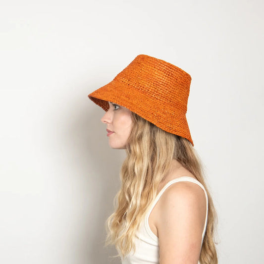 A woman with long, wavy blonde hair wears the WYETH TALI Raffia Bucket Hat in Rust and a white sleeveless top, standing in profile against a plain light background.